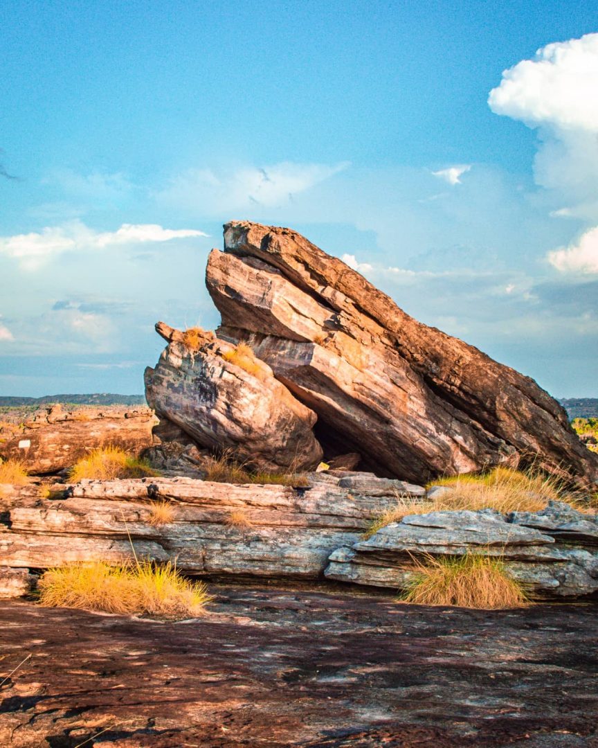 Layered sandstone boulders rise from the rocky Ubirr plateau beneath a bright blue sky. The rugged landscape is a scenic highlight of a Darwin to Kakadu weekend road trip.