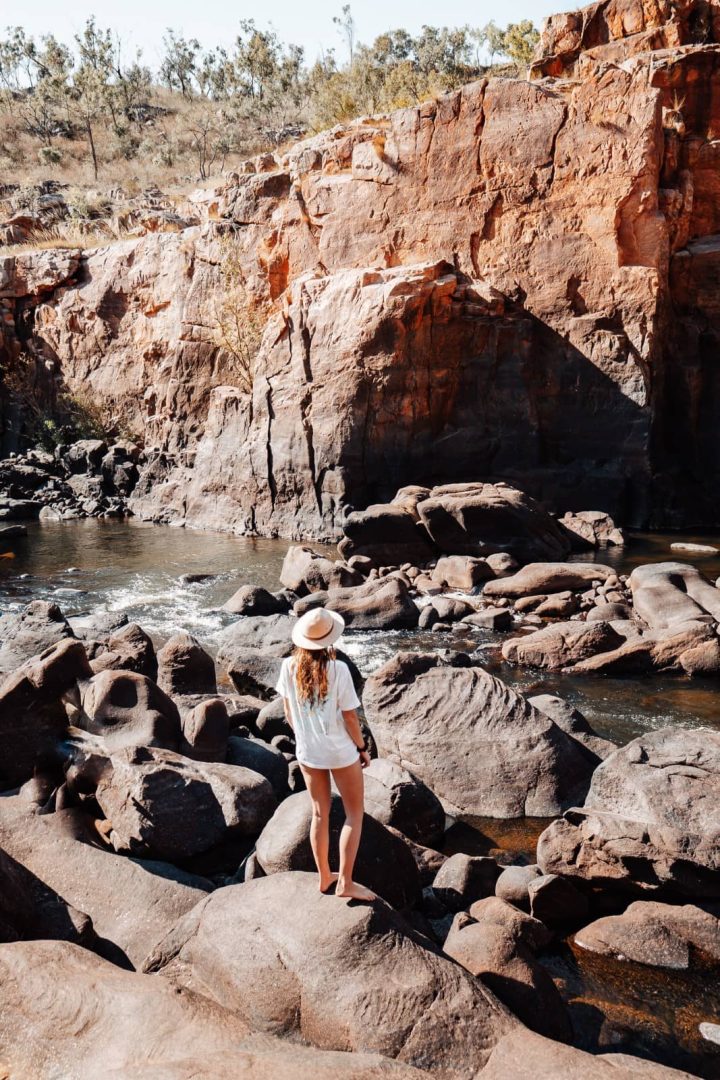 A woman stands on huge rounded rocks beside flowing water in Nitmiluk National Park. Rugged red cliffs rise behind the rocky section of Katherine Gorge.