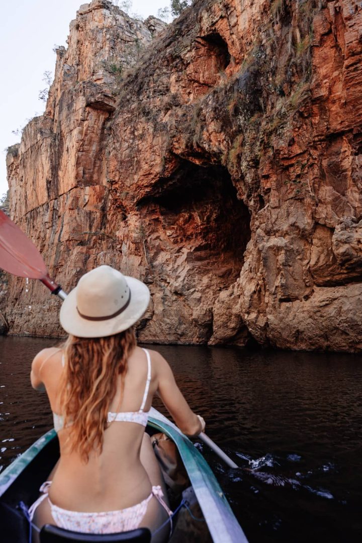 A woman paddles a canoe toward a dramatic hollow in the towering red cliffs of Katherine Gorge. The close cliffs show the scale of Nitmiluk from water level.