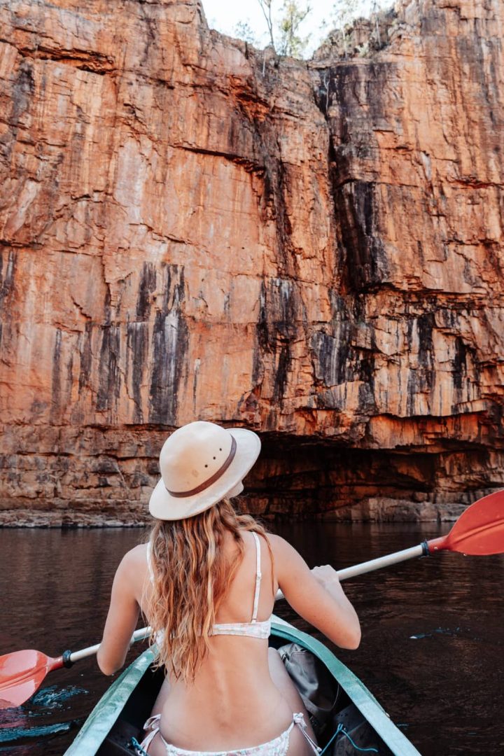 A woman in a sun hat paddles a canoe beneath an enormous orange sandstone wall in Katherine Gorge. Exploring Nitmiluk by canoe is a highlight of a Darwin to Katherine weekend road trip.