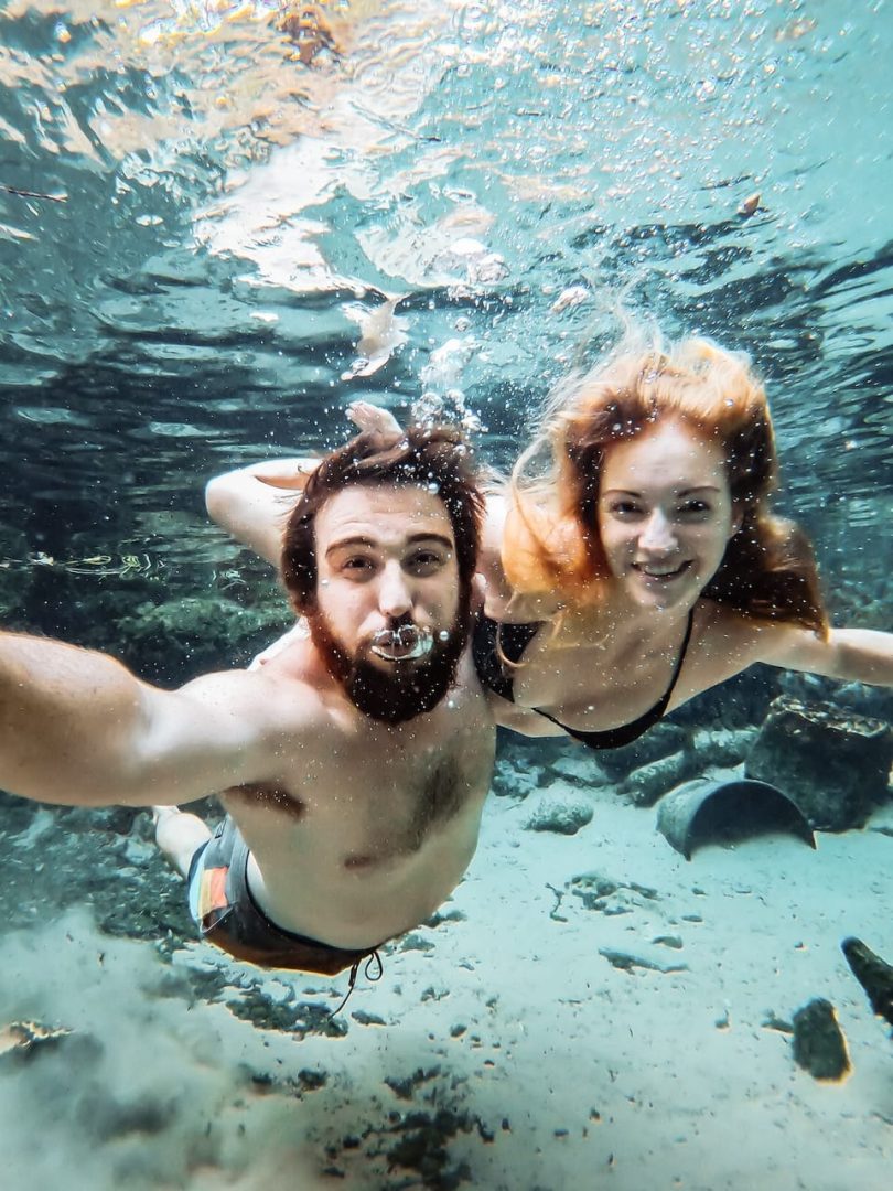 A man and woman swim underwater together in the clear blue water of Katherine Hot Springs. Bubbles surround them with rocks visible on the spring floor.