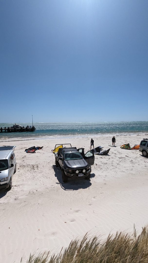 Four wheel drive vehicles and kitesurfing equipment sit on the white sand at Lancelin Beach beside turquoise water. Two people walk toward the ocean while a boat is moored at the end of a small jetty.