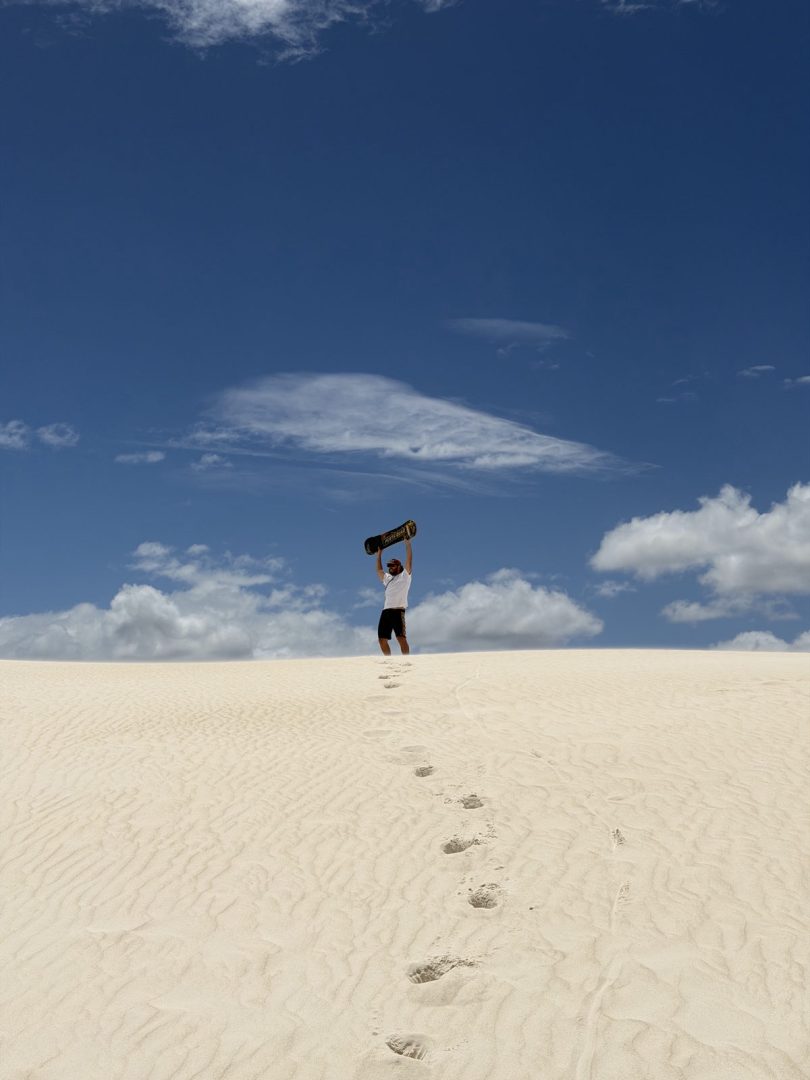 A man stands at the top of the bright white Lancelin sand dunes holding a sandboard above his head. His footprints lead up the dune beneath a deep blue sky during a Perth to Lancelin weekend.