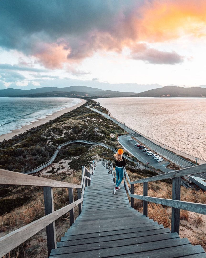A woman in an orange beanie walks down the wooden stairs at Truganini Lookout toward The Neck on Bruny Island. The narrow strip of land separates two sweeping beaches beneath dramatic sunset clouds during this Tasmania road trip from Devonport.