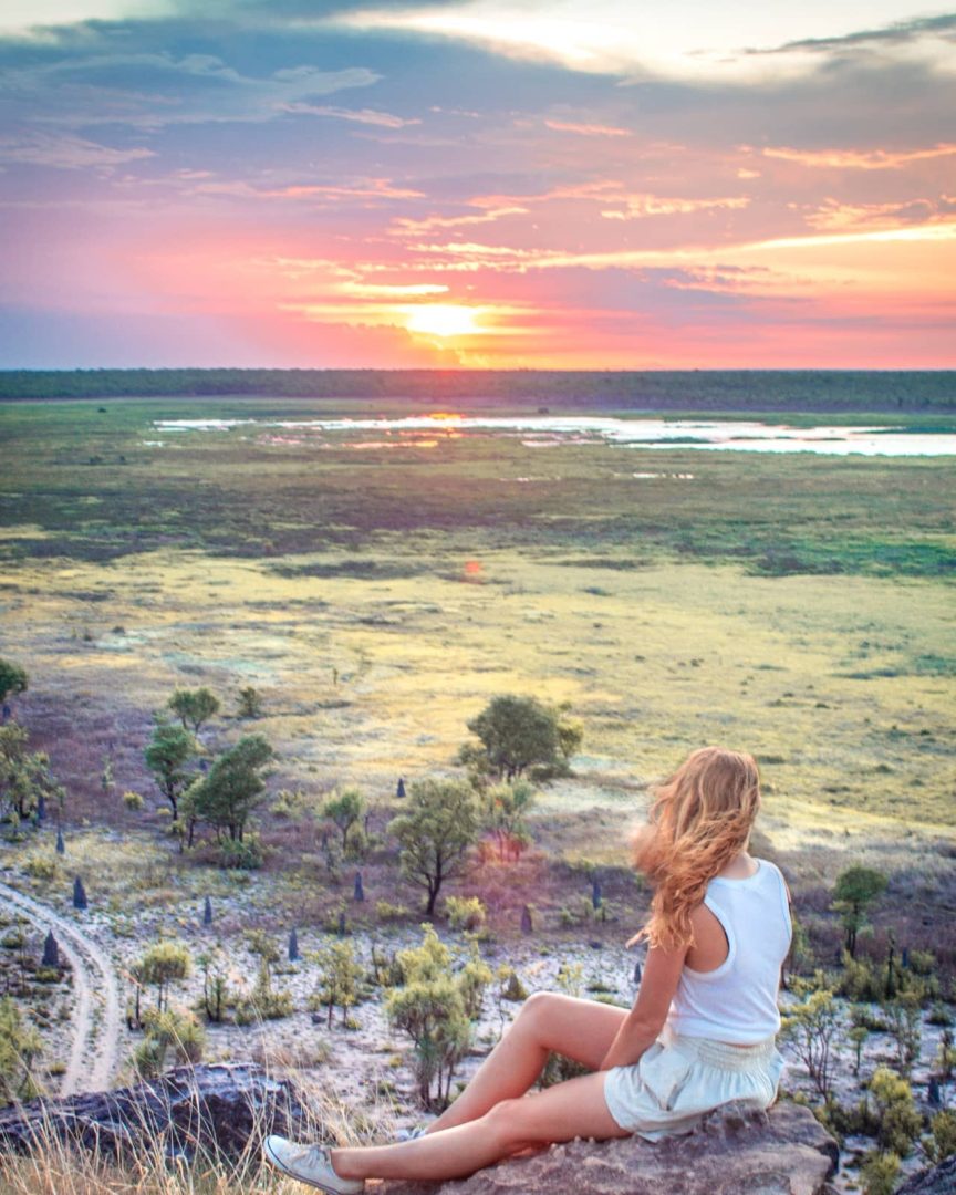 A woman sits on a rocky Ubirr lookout watching the sunset glow pink and orange above the Kakadu wetlands. The panoramic view is a memorable moment on a Darwin to Kakadu weekend road trip.