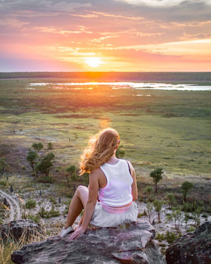 A woman sits on a rocky ledge at Ubirr while the sun sets over the expansive Kakadu wetlands. Golden light reflects across the floodplain beneath pink and orange clouds.
