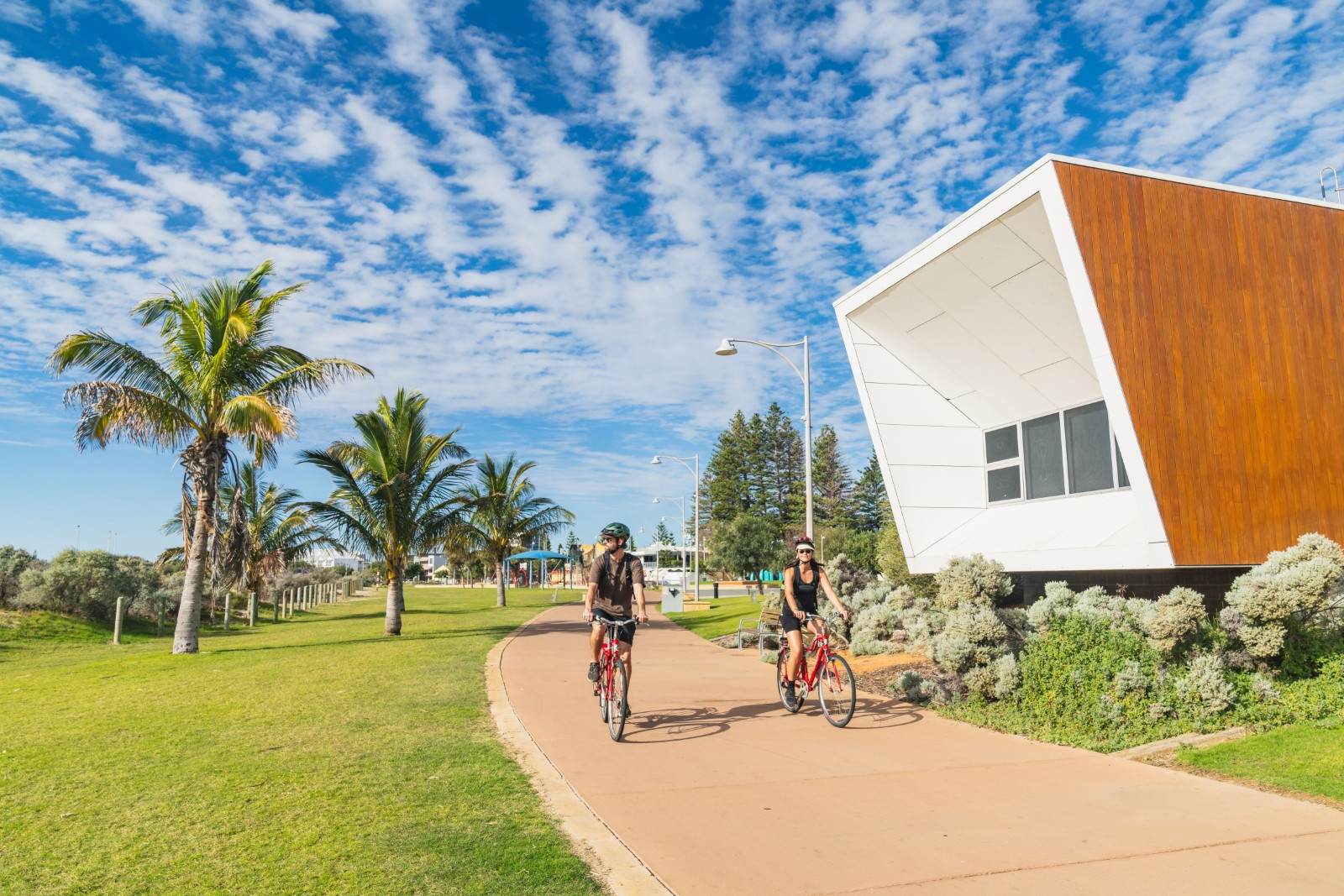 Two cyclists ride along the Geraldton Foreshore Walk beside palm trees and green lawns on a bright sunny day. Source. Tourism Western Australia.