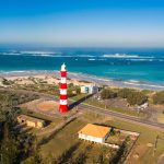 Aerial view of the red and white striped Point Moore Lighthouse in Geraldton with coastal dunes and the bright blue Indian Ocean behind it. Source. Tourism Western Australia.