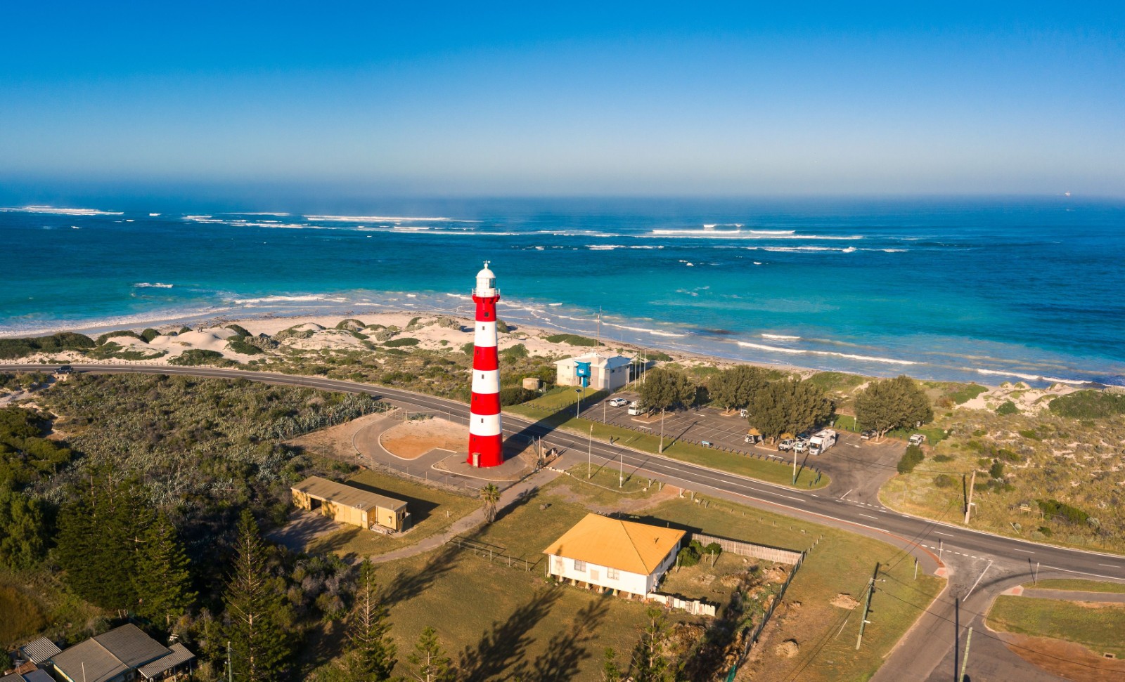 Aerial view of the red and white striped Point Moore Lighthouse in Geraldton with coastal dunes and the bright blue Indian Ocean behind it. Source. Tourism Western Australia.