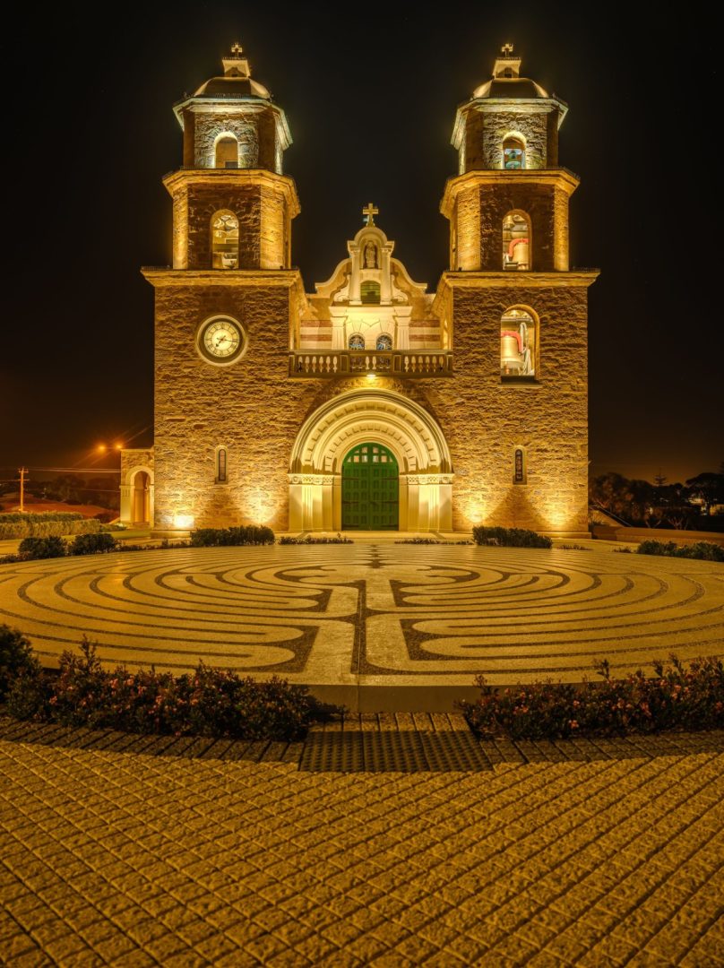 St Francis Xavier Cathedral in Geraldton illuminated in golden light at night with its twin towers rising above a large circular labyrinth in the courtyard. Source. Tourism Western Australia.