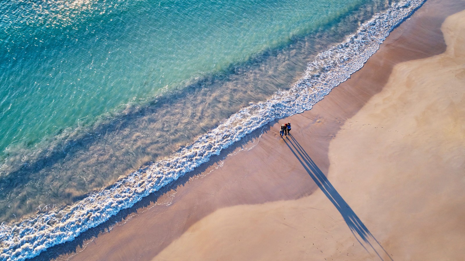 Aerial view of three people standing at the waters edge on Town Beach in Geraldton with clear turquoise water washing onto the sand. Their long shadows stretch across the beach in the low sunlight. Source. Tourism Western Australia.