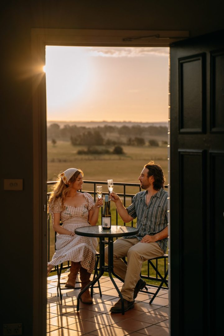 A couple toasting with drinks on their room balcony at sunset, looking over the paddocks at Camden Valley Inn.
