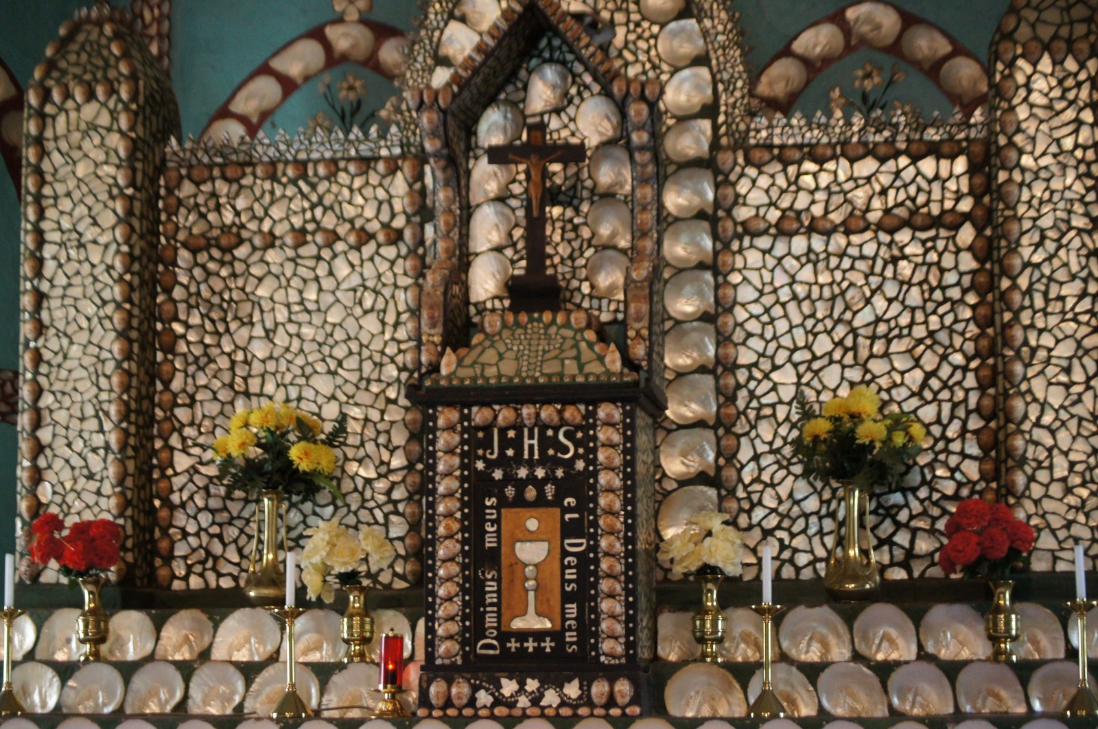 Mother of pearl altar inside Sacred Heart Church, Beagle Bay, Western Australia, decorated with shells, flowers and a wooden crucifix. The altar includes the text "IHS" and "Dominus meus et Deus meus".